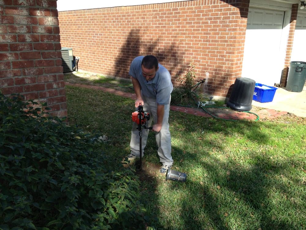 Pest control technician using a motorized auger to drill a hole for a subterranean termite bait station in a residential lawn.