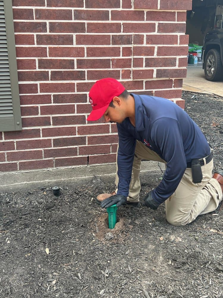 Licensed technician kneeling in a mulch bed beside a brick home, installing a green termite bait station in the soil.