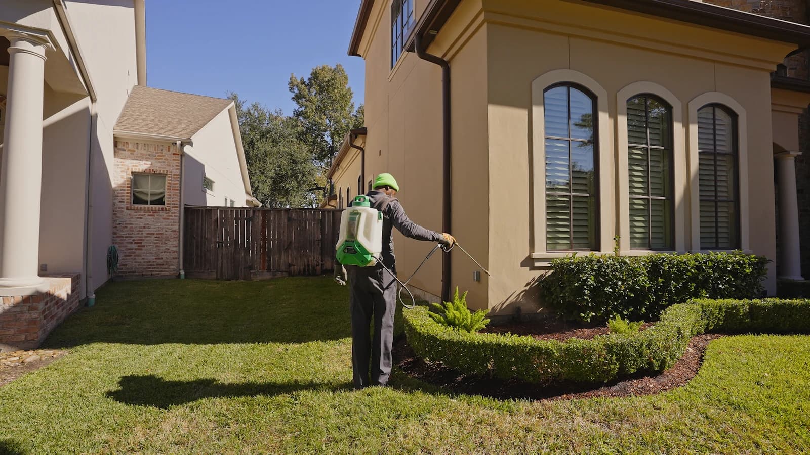 Pest control technician wearing a backpack sprayer and applying a liquid perimeter treatment along the foundation of a two-story home.