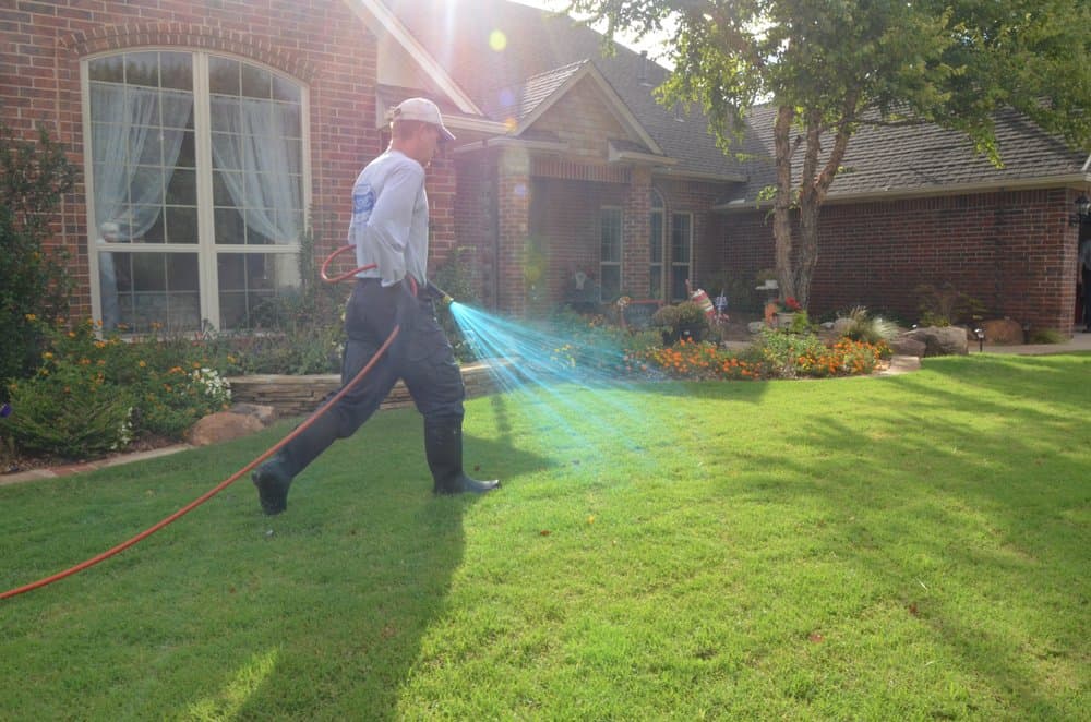 Technician in rubber boots walking a residential lawn and applying a spray treatment in front of a brick house.