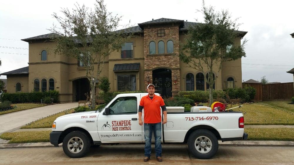 Stampede Pest Control technician in an orange polo and cap standing beside a branded white service truck with spray equipment, parked in front of a two-story home.